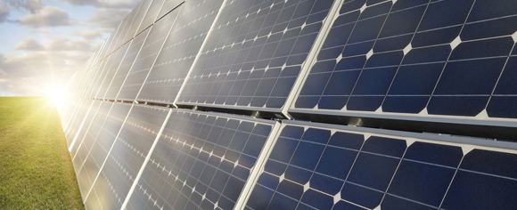 Rząd paneli słonecznych na zielonym polu, oświetlonych jasnym słońcem zza chmur.