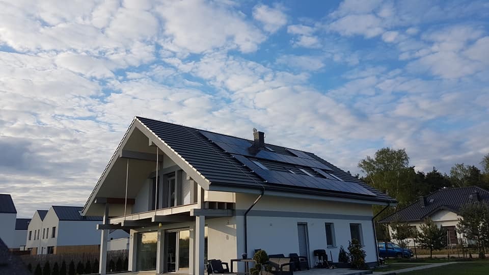 Dom jednorodzinny z zainstalowanymi panelami fotowoltaicznymi na dachu, widok z poziomu gruntu, na tle błękitnego nieba z chmurami.