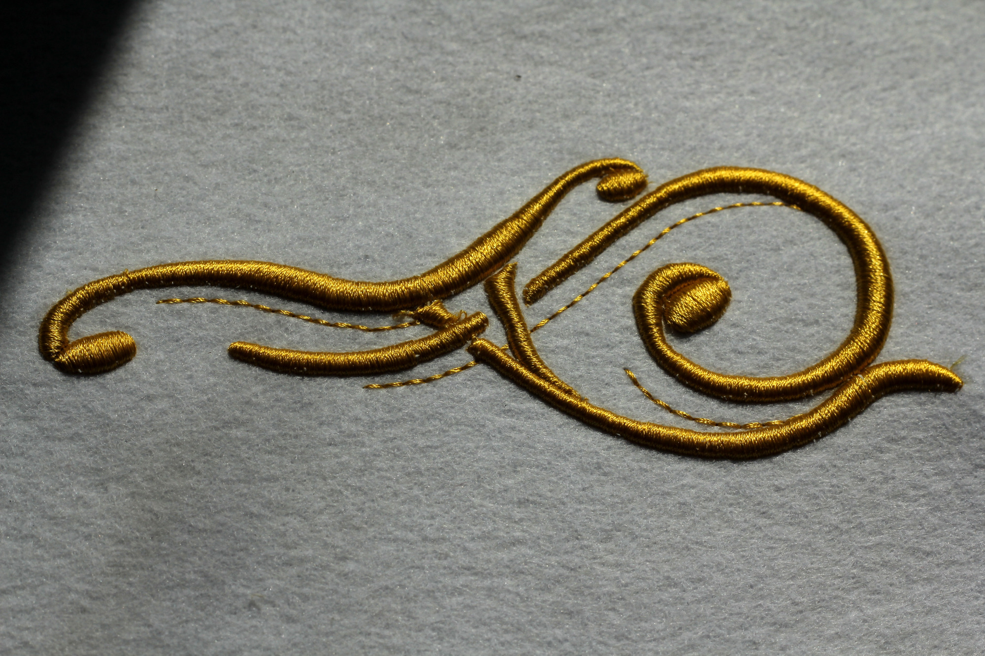 Detal haftu wypukłego w kolorze złotym na szarym filcu, przedstawiający stylizowany ornament roślinny.