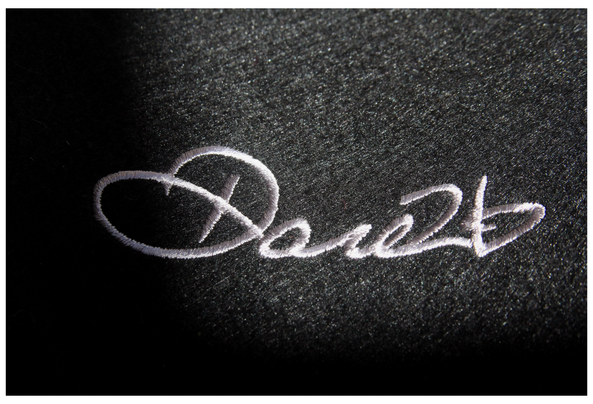 Biały, maszynowy napis 'Dare2b' na ciemnoszarym, teksturowanym materiale, oświetlony z góry, z widocznymi detalami ściegów.