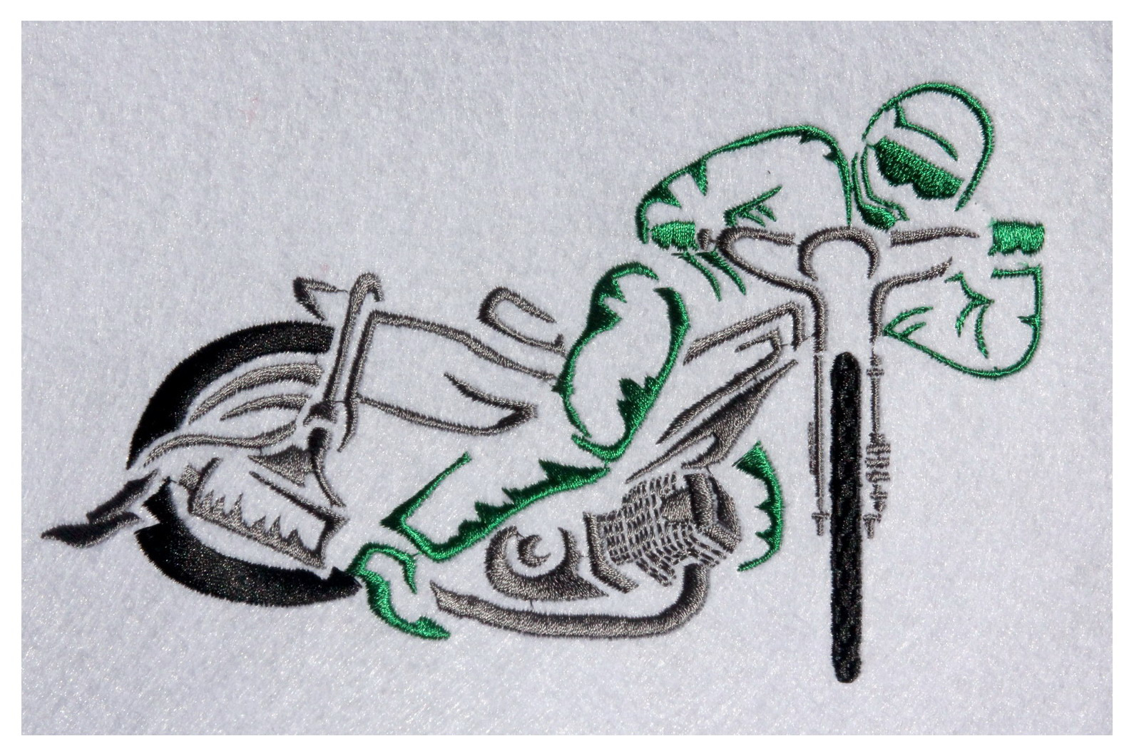 Wykonany z nici haft na białym materiale przedstawiający motocyklistę na motorze żużlowym z zielonymi akcentami.