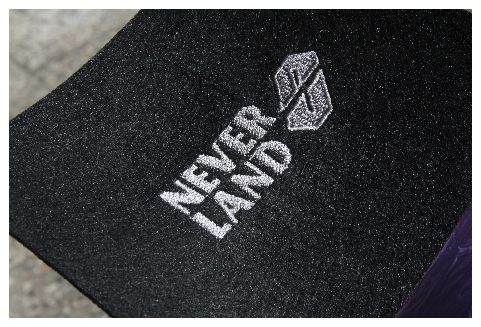 Czarny materiał z wyhaftowanym białym napisem 'Never Land' i graficznym logo powyżej, widoczne z bliska.