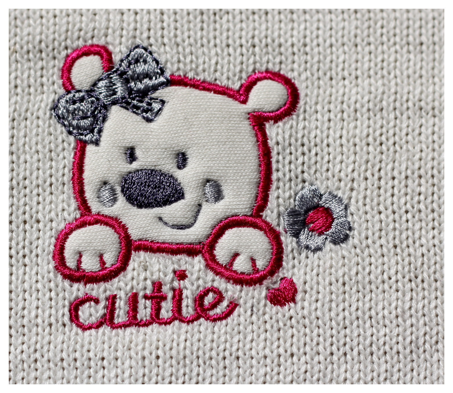 Haftowany wzór misia z kokardą i kwiatkiem na dzianinowej tkaninie w odcieniach bieli, różu i szarości z napisem 'cutie'.