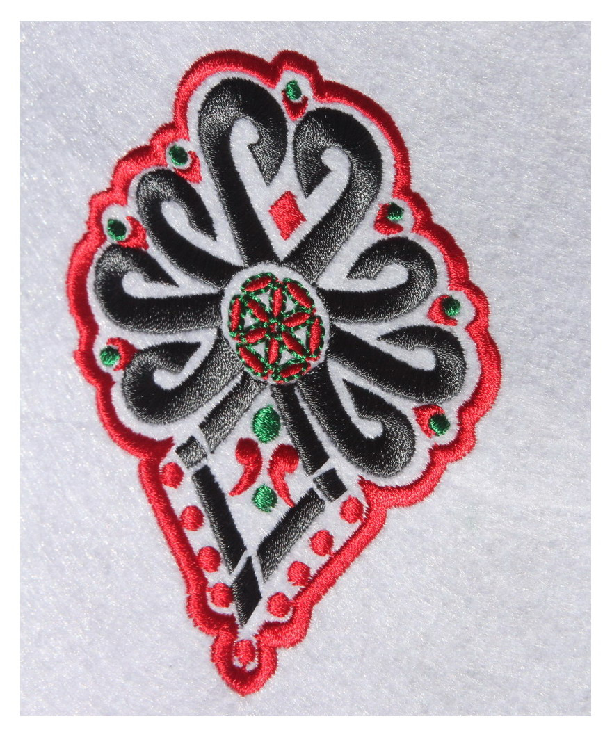 Ozdobny, haftowany wzór w kolorach czarnym, czerwonym i zielonym na białym tle, przypominający kwiat lub ornament.