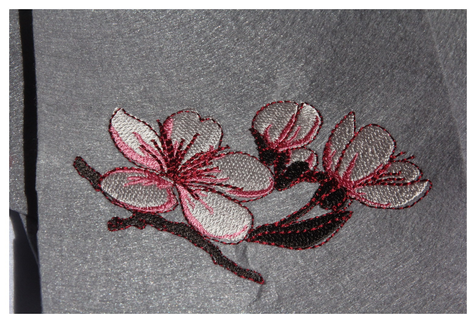 Delikatny haft przedstawiający gałązkę kwitnącej wiśni z trzema kwiatami, wykonany czerwoną i srebrną nicią na szarym, lekko połyskującym materiale.