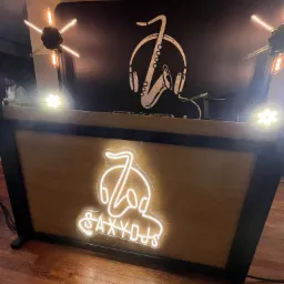 Podświetlane stanowisko DJ-a z neonowym logo 'Zoo & SAXXDJs' na drewnianym panelu, czarne tło z grafiką saksofonu w słuchawkach, oświetlone dekoracyjnymi lampami w kształcie gwiazd.