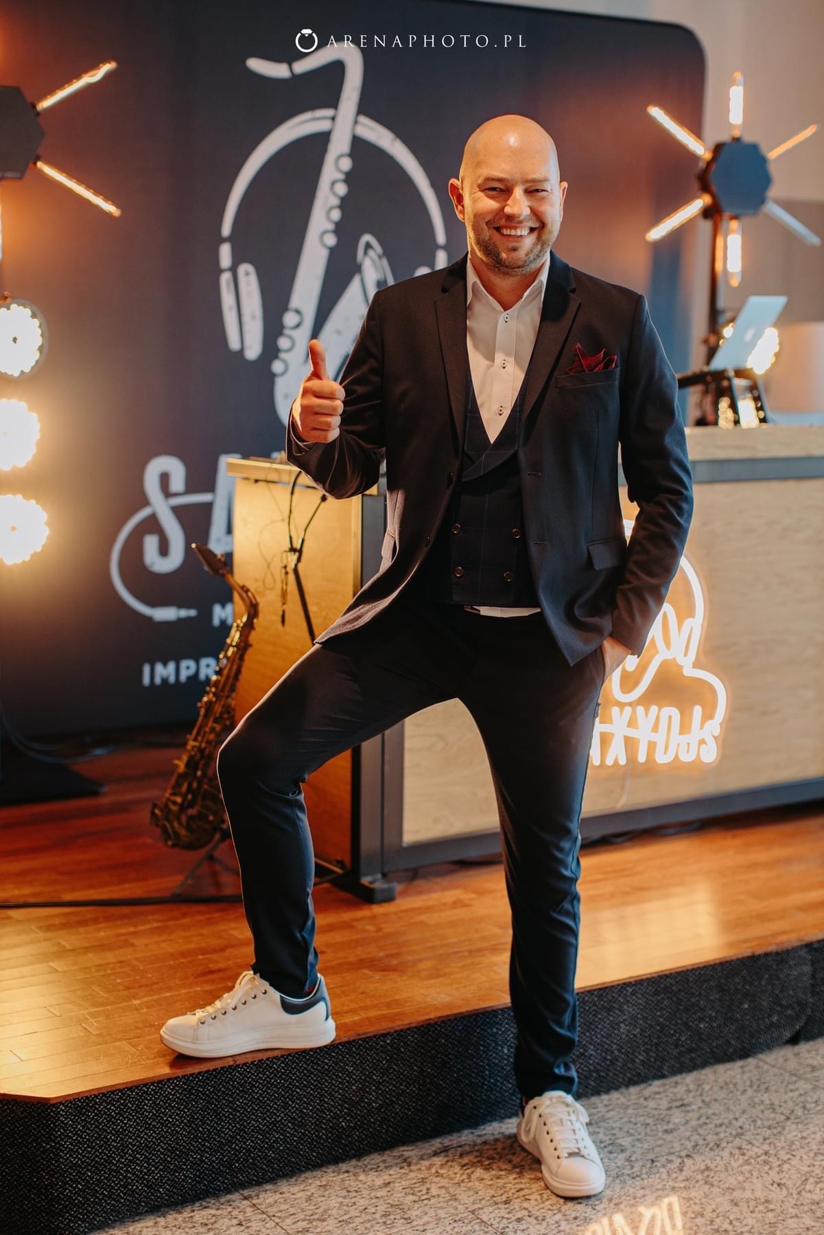 Uśmiechnięty mężczyzna w garniturze i białych sneakersach, stojący na scenie z saksofonem w tle, kciuk w górę.