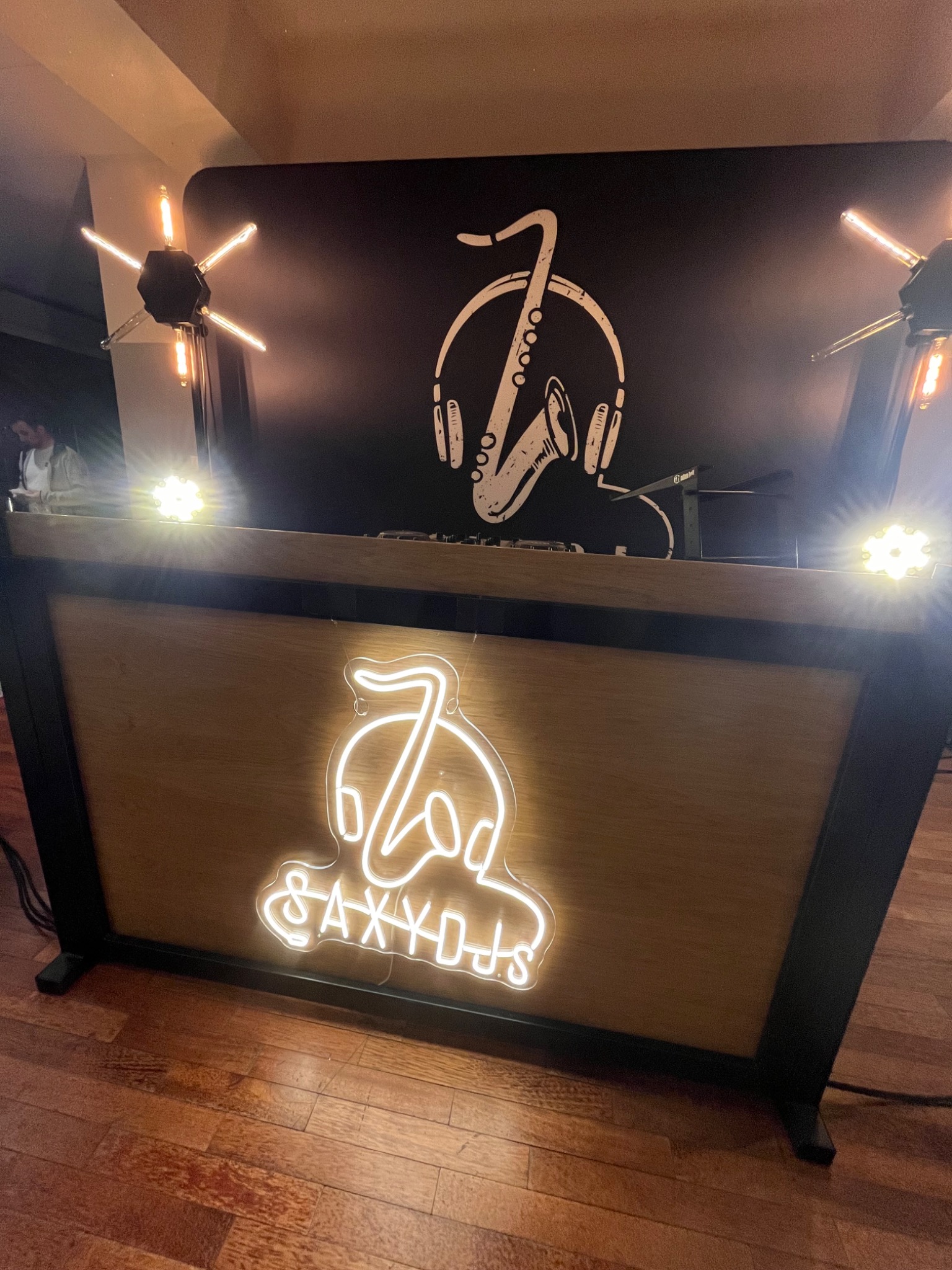 Podświetlane stanowisko DJ-a z neonowym logo 'Zoo & SAXXDJs' na drewnianym panelu, czarne tło z grafiką saksofonu w słuchawkach, oświetlone dekoracyjnymi lampami w kształcie gwiazd.