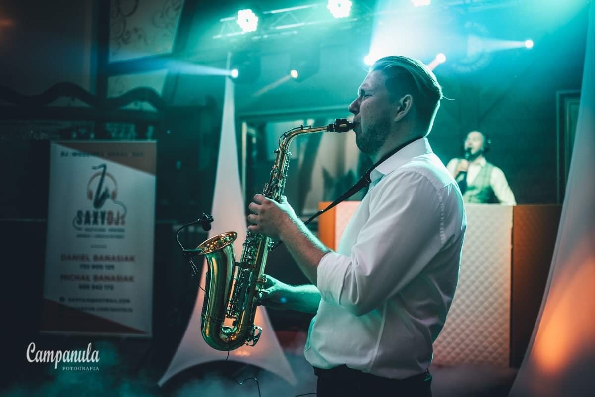 Saksofonista w białej koszuli gra na saksofonie podczas występu na scenie, w tle widać wokalistę przy pulpicie.
