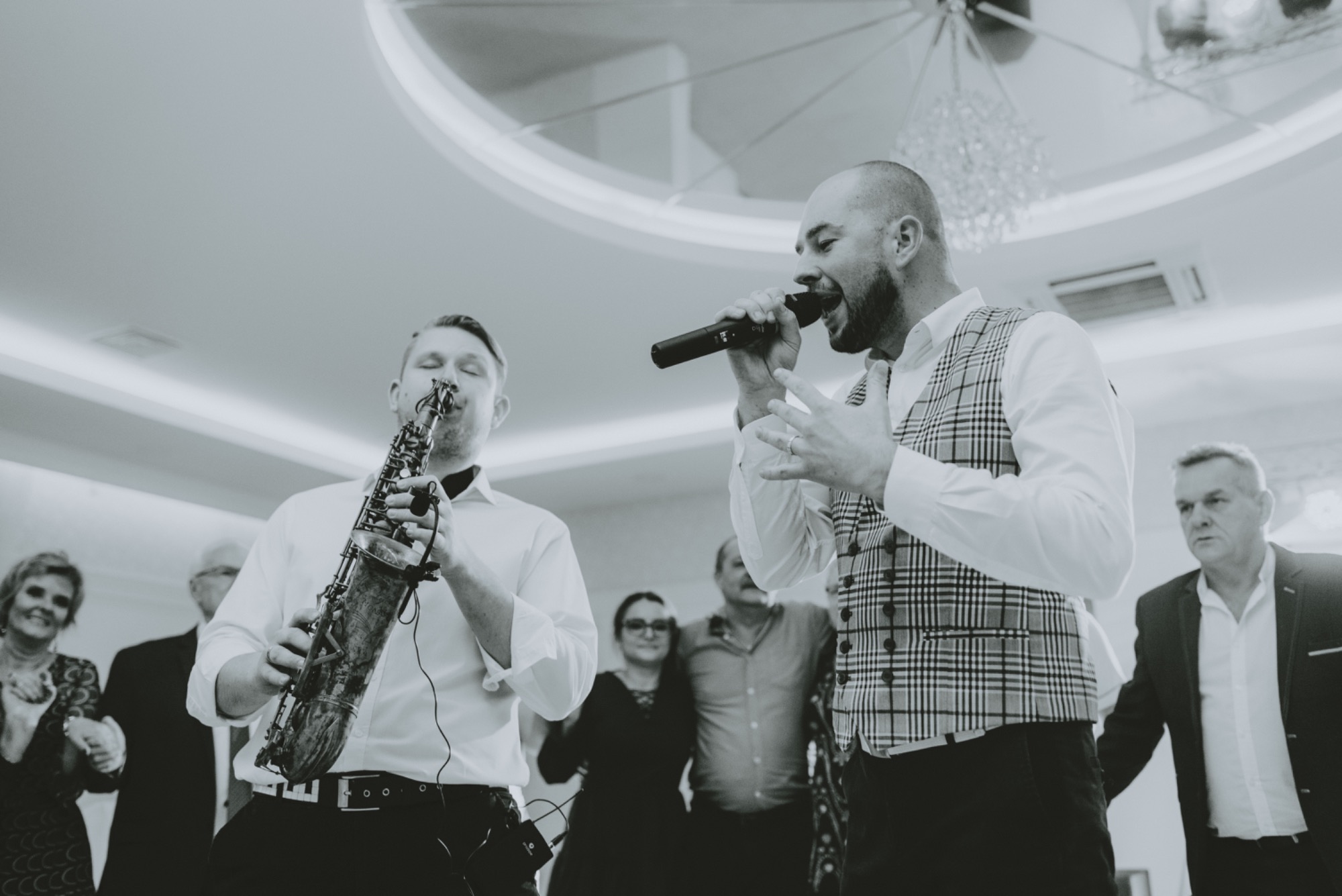 Wokalista z mikrofonem i saksofonista podczas występu na weselu, w tle goście i dekoracyjne oświetlenie sufitowe, zdjęcie w odcieniach szarości.