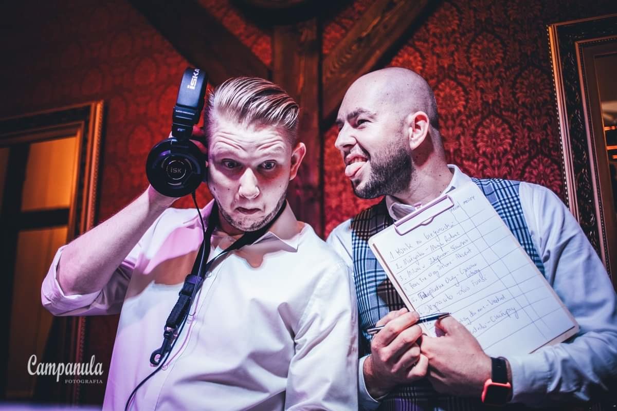 Dwóch mężczyzn pozuje: jeden z słuchawkami na uszach robi minę, drugi z wyciągniętym językiem trzyma listę utworów na kartce.