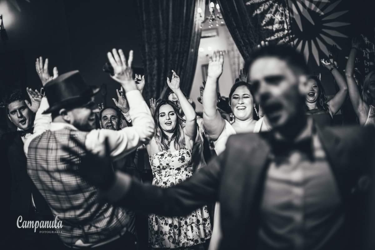 Czarno-białe zdjęcie z imprezy, na pierwszym planie rozmazana sylwetka mężczyzny w garniturze i muszce, w tle uśmiechnięci ludzie z podniesionymi rękami, w tym mężczyzna w kapeluszu i kraciastej...