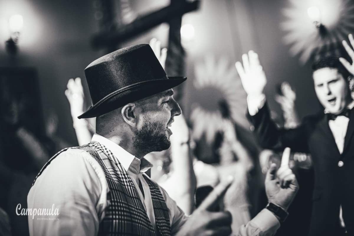 Czarno-białe zdjęcie mężczyzny w cylindrze i kraciastej kamizelce, śpiewającego z uśmiechem na twarzy, w tle rozmazane postacie z podniesionymi rękami podczas występu.