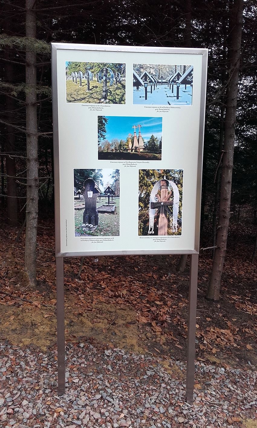 Tablica informacyjna z fotografiami cmentarzy wojennych, umieszczona na zewnątrz, w otoczeniu drzew i opadłych liści.