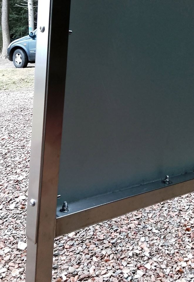 Metalowa konstrukcja ramy wystawienniczej z szarą płytą, widoczne śruby mocujące na tle żwirowej nawierzchni i fragmentu samochodu.