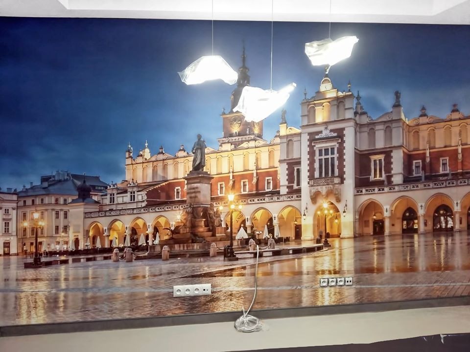 Tapeta winylowa z nadrukiem Rynku Głównego w Krakowie, widoczne gniazdka elektryczne na dole tapety, oświetlenie sufitowe.