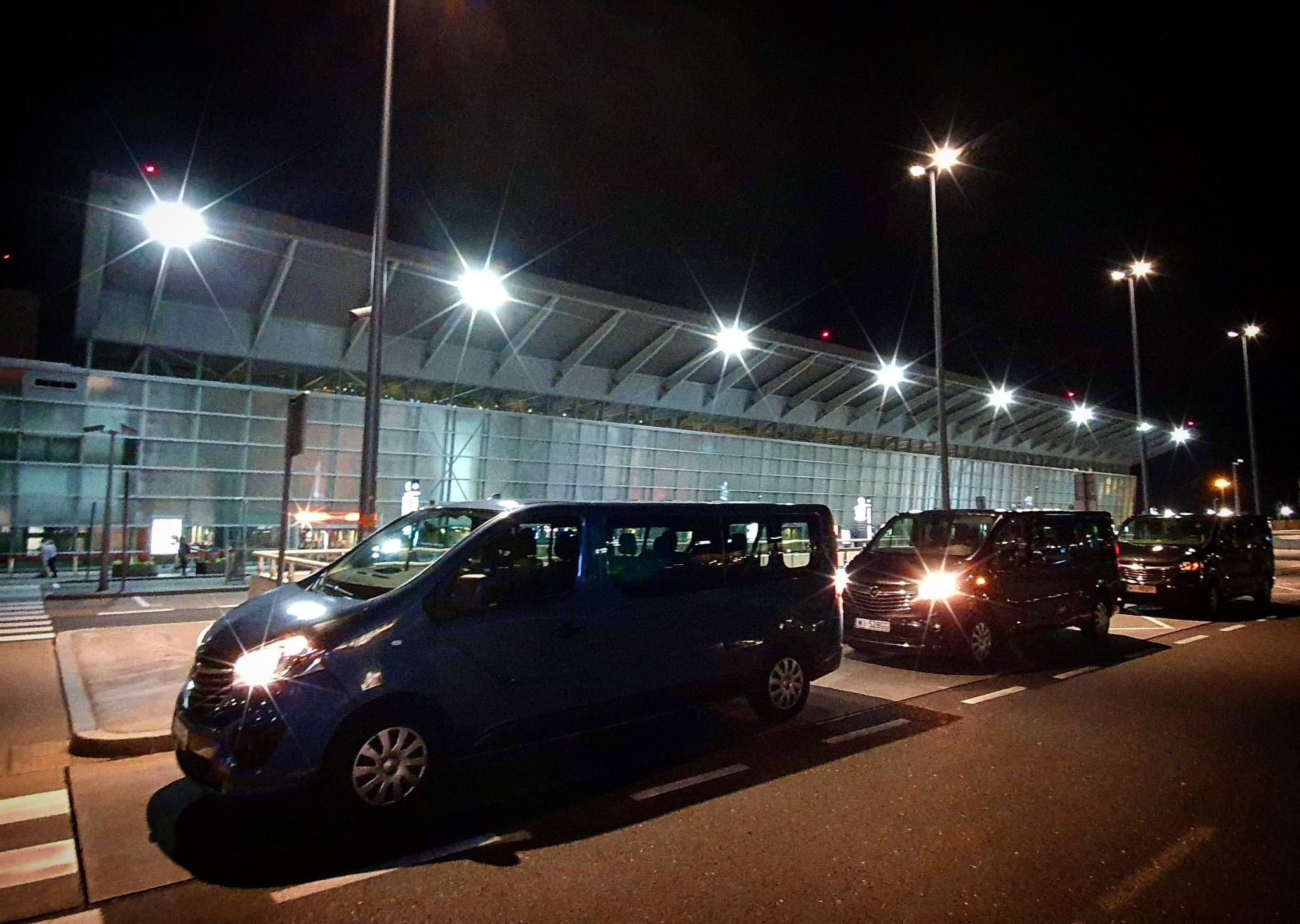 Nocny widok na rząd zaparkowanych busów przed budynkiem lotniska, oświetlonych latarniami, z widocznymi światłami samochodów.