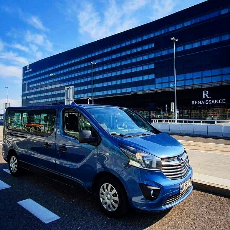 Niebieski bus Opel Vivaro zaparkowany na tle nowoczesnego budynku hotelu Renaissance z widocznym logo, błękitne niebo z chmurami w tle.