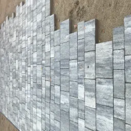 Fragment ścieżki z szarej kostki brukowej o nieregularnym kształcie, widoczny piasek jako podłoże, etap prac przed zakończeniem.