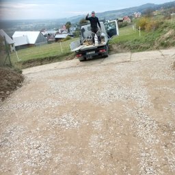 NORNIK PRACE BUDOWLANE ORAZ ROBOTY ZIEMNE - Przygotowany podjazd z tłucznia na posesji, pracownik na pace samochodu dostawczego pokazuje kciuk w górę, w tle zabudowania wiejskie i wzgórza.