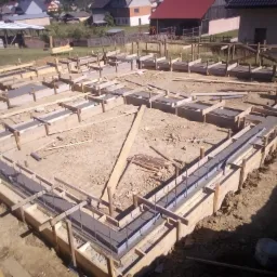 Wylane fundamenty zbrojone stalą, drewniane szalunki w ziemi, widok z góry na plac budowy z domami w tle.