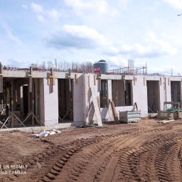 NORNIK PRACE BUDOWLANE ORAZ ROBOTY ZIEMNE - Wznoszenie ścian parterowego domu z betonu komórkowego, widoczne zbrojenia, stemple budowlane i ślady opon na gruncie.