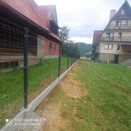NORNIK PRACE BUDOWLANE ORAZ ROBOTY ZIEMNE - Nowo postawione ogrodzenie panelowe z podmurówką betonową, oddzielające posesje z domami w stylu góralskim, na tle zalesionych wzgórz.