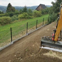 NORNIK PRACE BUDOWLANE ORAZ ROBOTY ZIEMNE - Żółta koparka przygotowuje teren pod montaż ogrodzenia panelowego z podmurówką betonową na zboczu z widokiem na dom i zalesione wzgórza w tle.