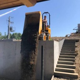 NORNIK PRACE BUDOWLANE ORAZ ROBOTY ZIEMNE - Żółta wywrotka z operatorem wysypuje ziemię obok betonowego muru oporowego i schodów terenowych na placu budowy w słoneczny dzień.