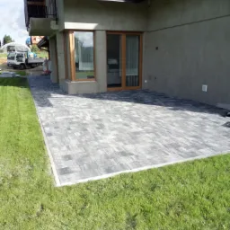 Szara kostka brukowa ułożona na tarasie przylegającym do domu z drewnianą stolarką okienną, widok z poziomu trawnika.