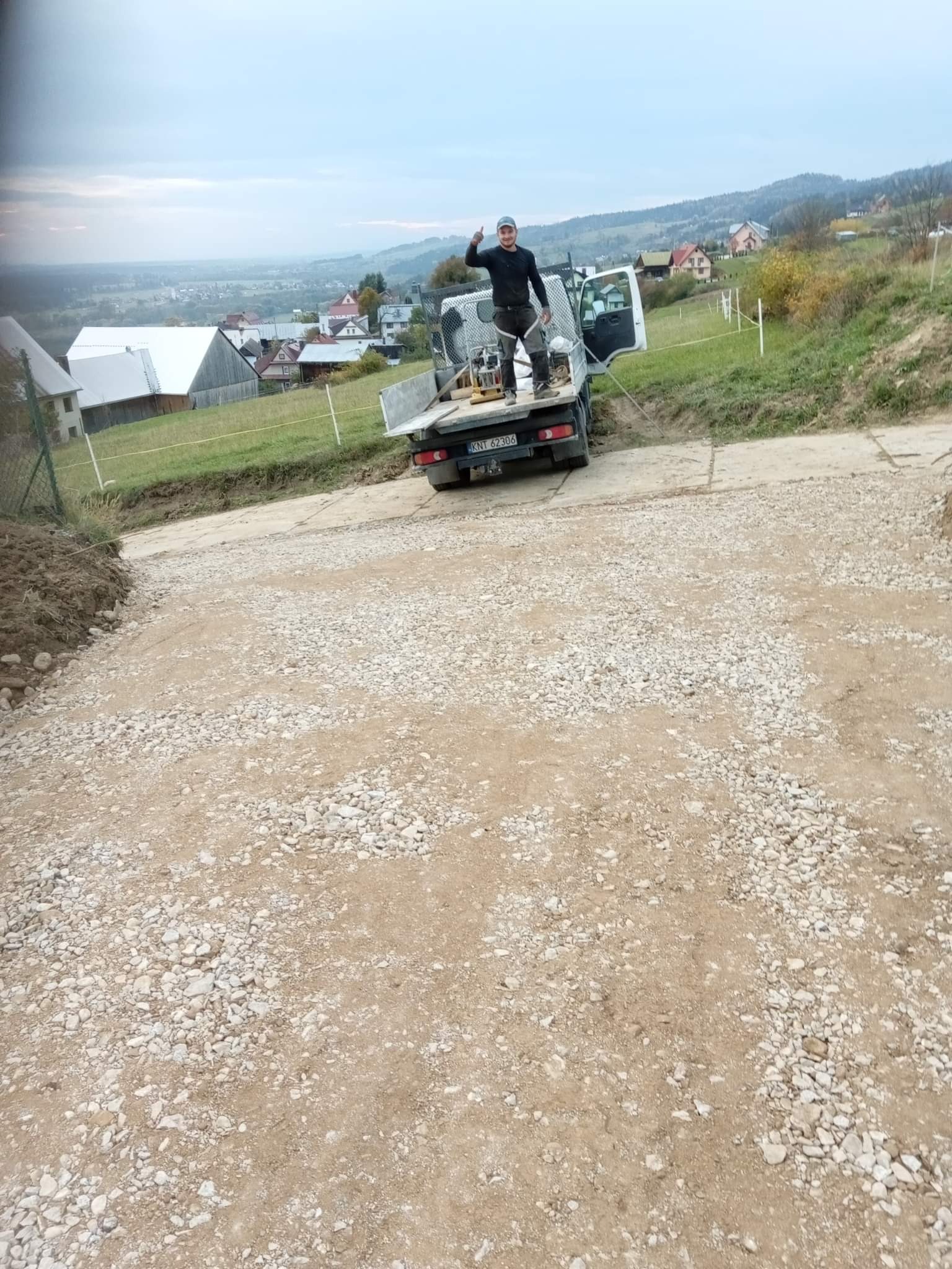 Przygotowany podjazd z tłucznia na posesji, pracownik na pace samochodu dostawczego pokazuje kciuk w górę, w tle zabudowania wiejskie i wzgórza.