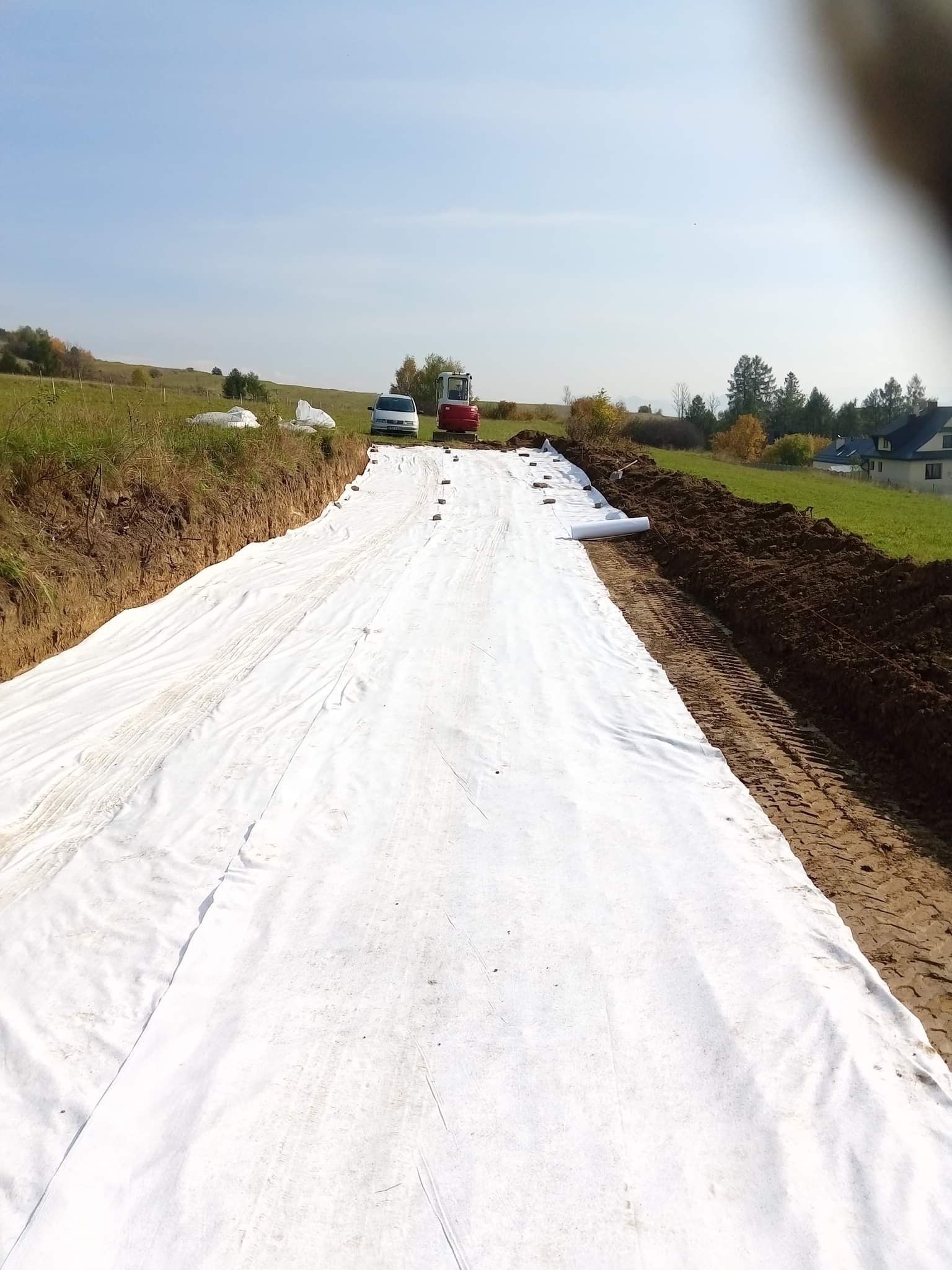 Przygotowanie terenu: Wykop wyłożony białą geowłókniną, zabezpieczony kamieniami, z widocznym minikoparką i samochodem w tle, na tle zielonego krajobrazu.