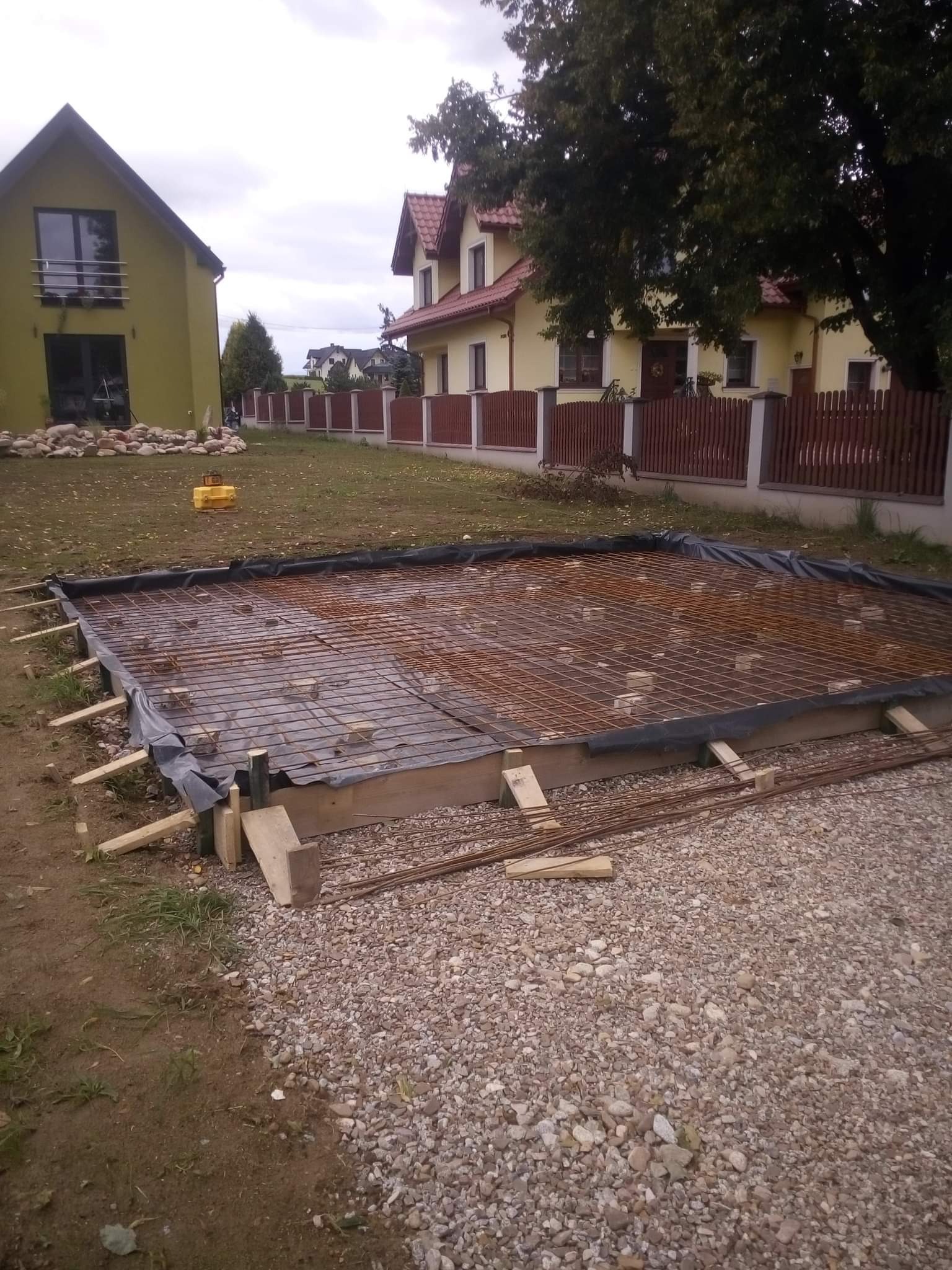 Przygotowanie zbrojenia fundamentu pod budowę domu jednorodzinnego, widoczne drewniane szalunki, siatka zbrojeniowa na folii, w tle domy i drzewa.