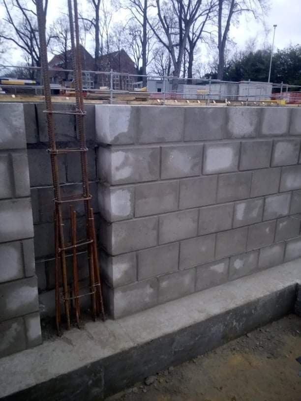 Wznoszenie ściany fundamentowej z szarych bloczków betonowych, z widocznym zbrojeniem pionowym i poziomym, na tle budynku w budowie i drzew bez liści.