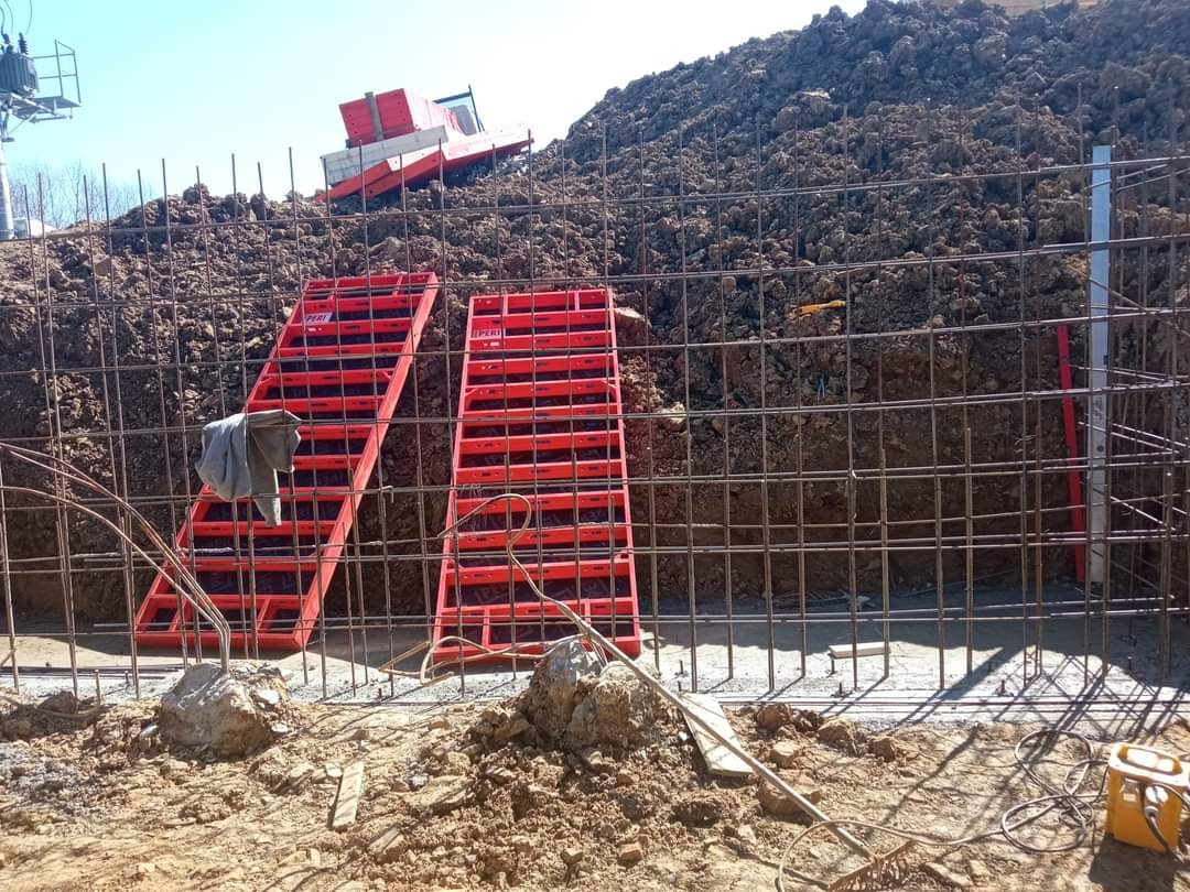 Zbrojenie wykopu pod fundamenty z użyciem czerwonych szalunków PERI, na zboczu widoczna koparka, przewody elektryczne i narzędzia na ziemi.