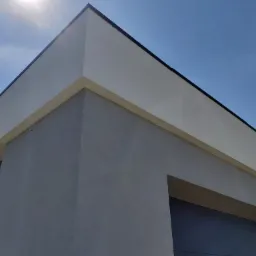 Narożnik budynku z nowoczesną elewacją w odcieniach szarości i bieli, widok z dołu na tle błękitnego nieba, z widocznym kablem zwisającym ze ściany.