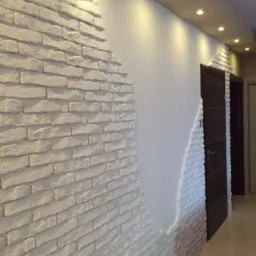 Wykończenie wnętrza z częściowo ułożoną dekoracyjną białą cegłą na ścianie, widoczne halogenowe oświetlenie sufitowe i fragmenty drzwi.