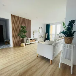 Jasne wnętrze salonu z drewnianą ścianą z lameli, białym regałem, kanapą i roślinami w doniczkach, podkreślone parkietem.