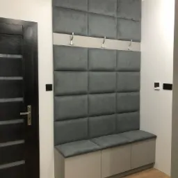 Szara tapicerowana ściana z wieszakami i siedziskiem w przedpokoju, widoczna obok ciemna drewniana drzwi.