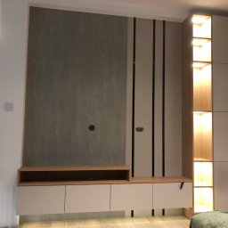 FIN-DREW CIĘŻCY KLAUDIA CIĘŻKA - Nowoczesna zabudowa ściany w salonie: szara ściana z otworem na kable, szafki z jasnego drewna z półką i szufladami, wysoka szafa z podświetlanymi półkami.