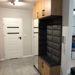 FIN-DREW CIĘŻCY KLAUDIA CIĘŻKA - Wysoka szafa z siedziskiem i tapicerowanym panelem ściennym w przedpokoju, drewniane fronty z czarnymi uchwytami, lustro na boku szafy.