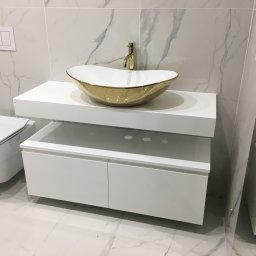 FIN-DREW CIĘŻCY KLAUDIA CIĘŻKA - Nowoczesna łazienka z białą szafką pod umywalkę, złotą umywalką w kształcie łodzi i złotą baterią, marmurowe płytki na ścianach i podłodze, wisząca toaleta po lewej stronie.