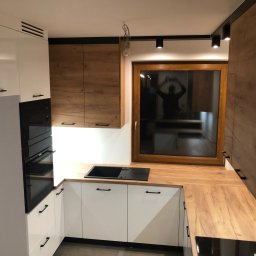 FIN-DREW CIĘŻCY KLAUDIA CIĘŻKA - Nowoczesna kuchnia w odcieniach bieli i drewna z czarnymi akcentami, widoczny fragment blatu drewnianego, czarny zlew i płyta indukcyjna, oświetlenie punktowe na suficie.