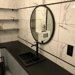 Nowoczesna łazienka z czarnym blatem, umywalką i okrągłym lustrem, marmurowe płytki na ścianach, minimalistyczna figurka odbijająca się w lustrze.