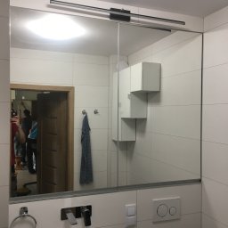 FIN-DREW CIĘŻCY KLAUDIA CIĘŻKA - Nowoczesna łazienka z białą armaturą, lustrem z oświetleniem LED i geometrycznymi szafkami wiszącymi; widoczne odbicie ekipy montażowej w lustrze.