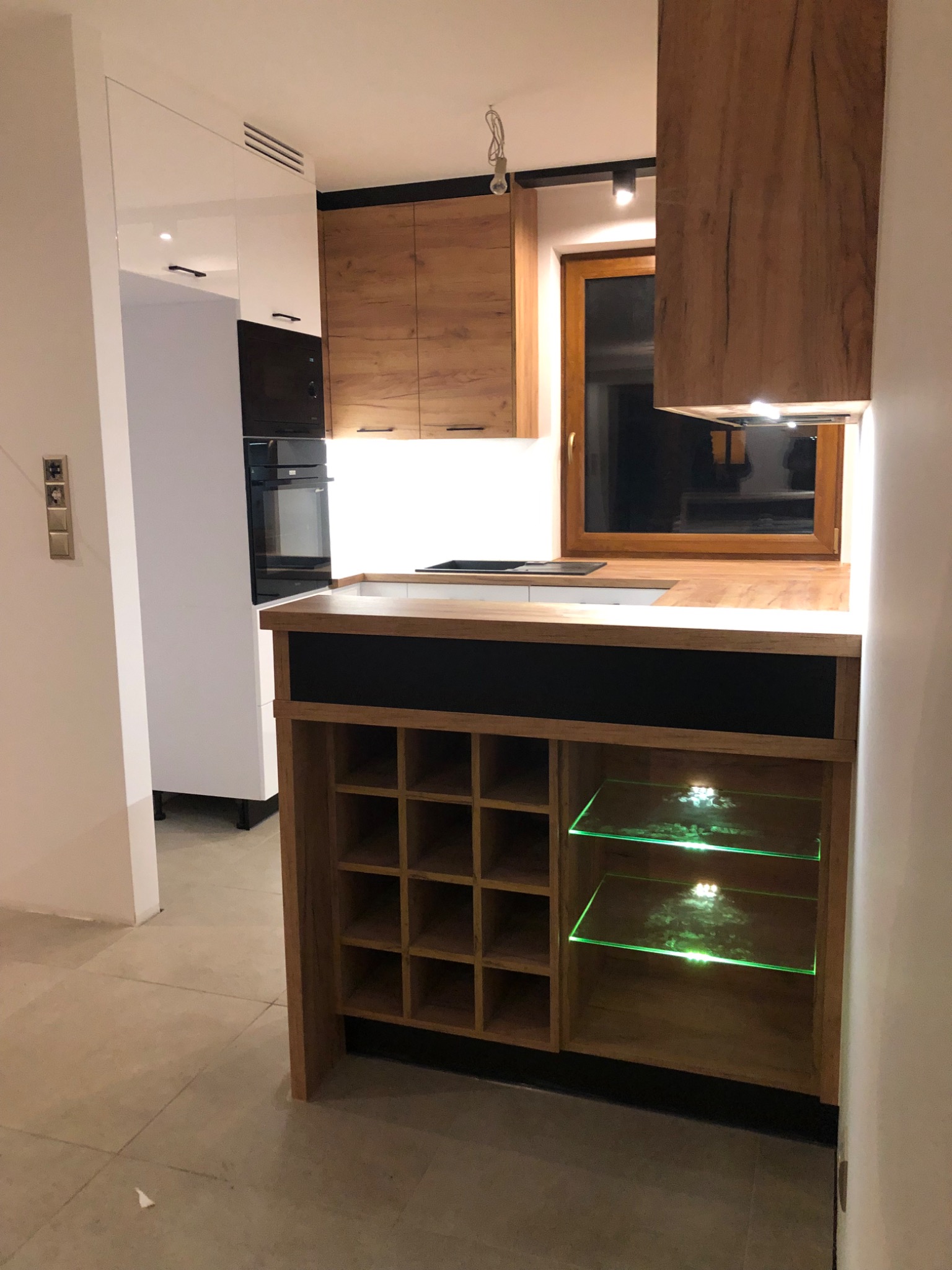 Nowoczesna kuchnia z drewnianymi szafkami, blatem i podświetlanymi półkami, z widocznym regałem na wino i oknem w tle.