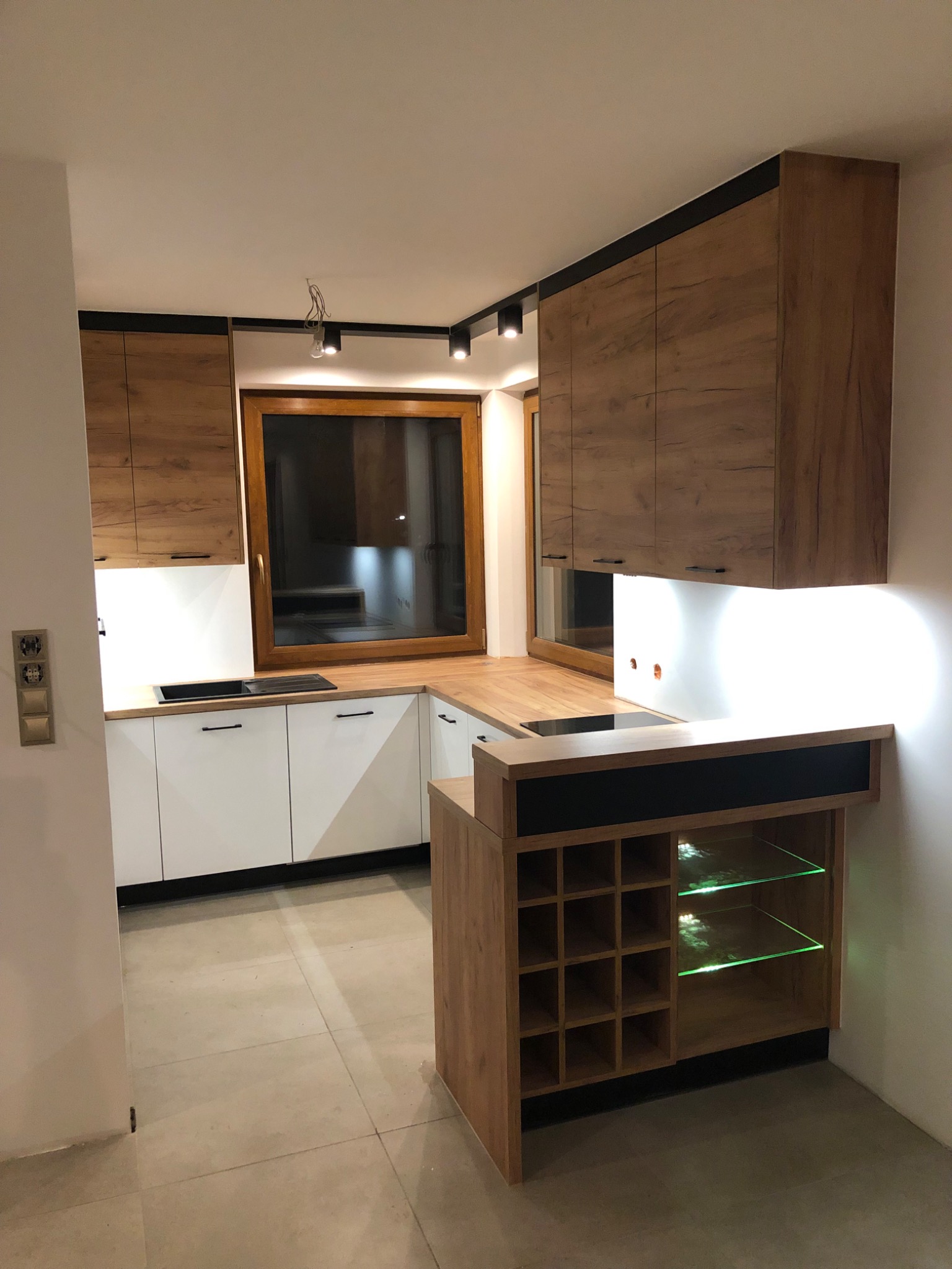 Nowoczesna kuchnia z białymi szafkami dolnymi, drewnopodobnymi szafkami górnymi z czarnymi akcentami i podświetlanym barkiem z półkami na wino.