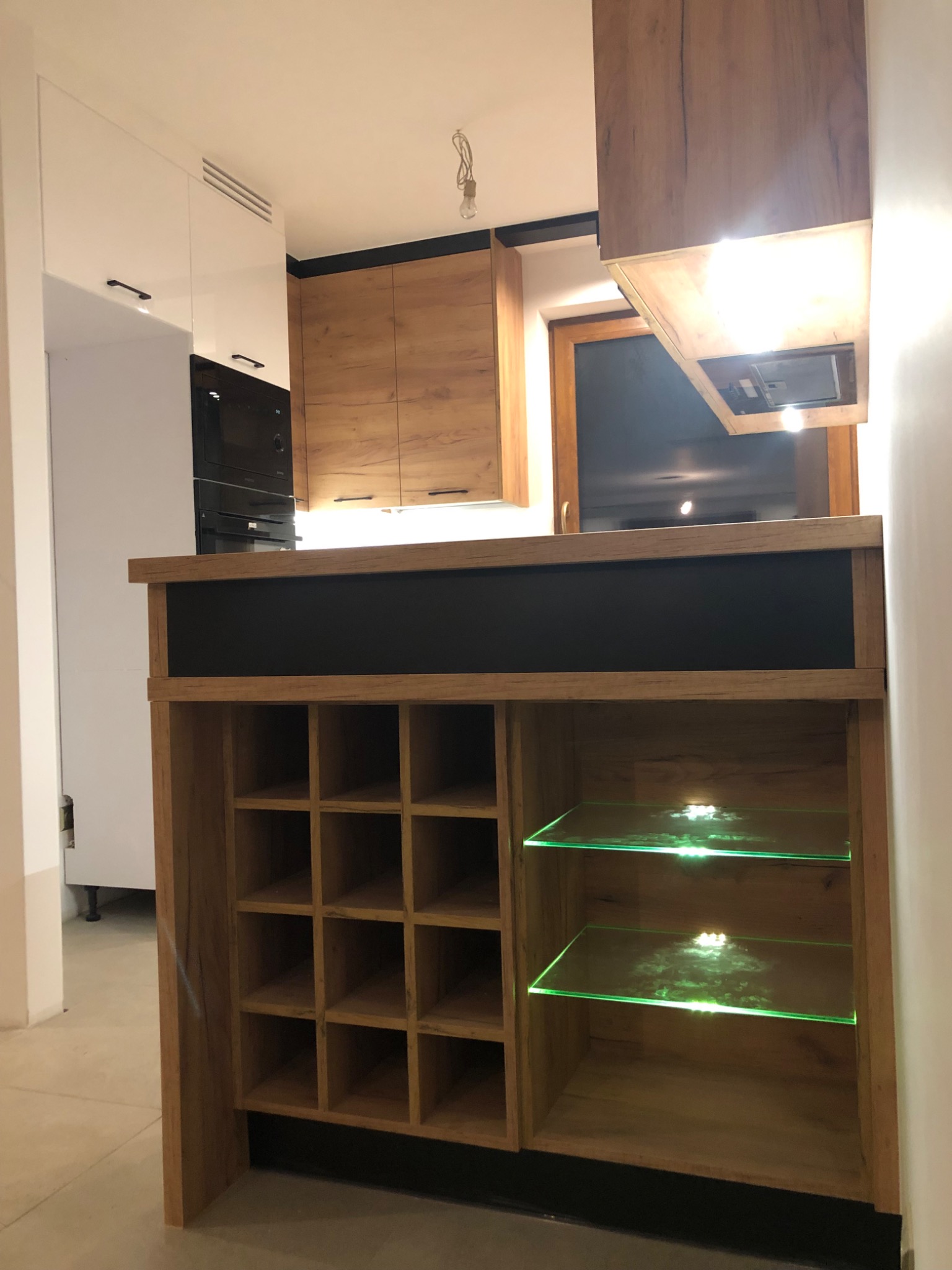 Nowoczesna kuchnia z drewnianymi frontami i wyspą z półkami na wino oraz podświetlanymi szklanymi półkami, widok na zabudowę piekarnika i okap.