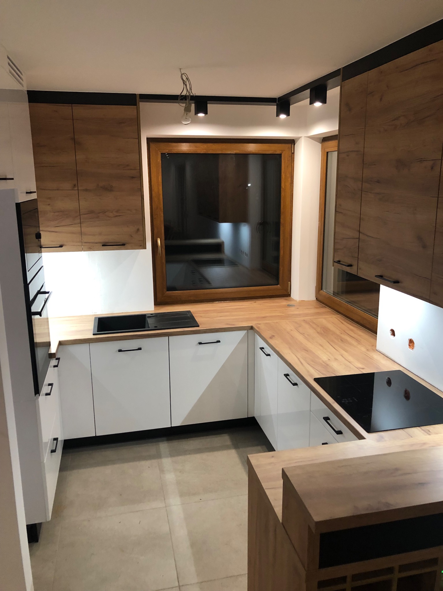 Nowoczesna kuchnia: białe szafki dolne z czarnymi uchwytami, górne szafki w odcieniach drewna, drewniany blat, czarny zlew i płyta indukcyjna, oświetlenie szynowe.