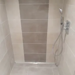 Nowoczesny prysznic z beżowymi i brązowymi płytkami, deszczownicą i odpływem liniowym w podłodze.